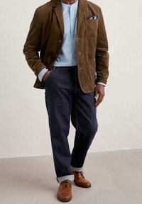 Brązowa marynarka z korduroyu, jasnoniebieska koszula z guzikami, ciemnoniebieskie dżinsy, brązowe skórzane buty oraz poszetka w niebieskie wzory.