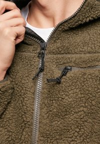 Giacca in pile verde oliva, con superficie texturizzata, chiusura con zip, cordino regolabile e una piccola tasca con zip.