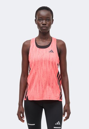 Žena oblečená v jasne ružovom tielku Adidas bez rukávov a čiernych legínach Adidas, stojaca pred jednoduchým svetlým pozadím.