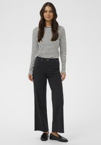 Femme avec de longs cheveux noirs portant un haut à rayures noir et blanc à manches longues, un pantalon noir à larges jambes taille haute et des mocassins noirs.