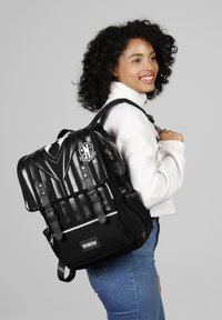 Le sac à dos présente un corps en toile noire avec un rabat rayé brillant, des sangles avant et un logo ; il comprend des fermetures éclair et une poche extérieure.