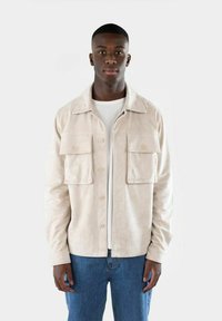 Giacca in suede beige con due tasche frontali, chiusura a bottone e colletto classico, abbinata a una t-shirt bianca e jeans blu.
