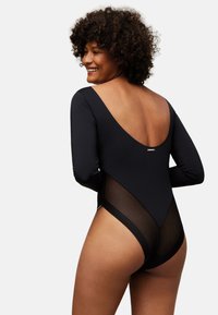 Body negro de manga larga con espalda baja, con paneles laterales de malla transparente y textura suave. Diseño minimalista con detalle de logo.