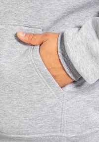 Grauer Sweatshirt mit weicher Texture, ausgestattet mit einer vorderen Tasche, die durch eine Naht umrandet ist. Die Bündchen sind gerippt, und eine Hand ruht in der Tasche.