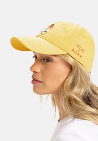 Yellow cotton baseball cap with an embroidered design featuring a mountain and sun, and "ATLAS x ROXY" logo in red on the side.