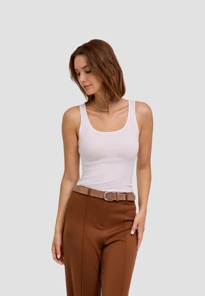 Jonge vrouw met schouderlang bruin haar, een wit hemdje, een bruine broek en een beige riem, kijkt naar beneden links.