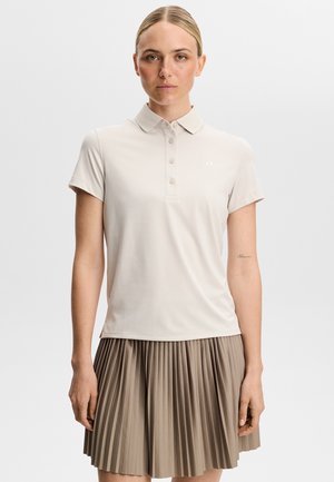 Kvinde iført en lys beige kortærmet polo og en taupe plisseret nederdel, stående mod en ensfarvet hvid baggrund.