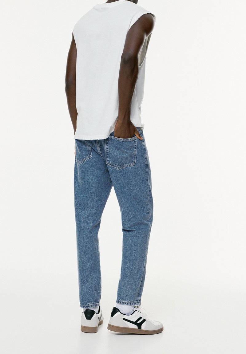 Light grey sleeveless top, blue denim jeans with a relaxed fit, and white sneakers with black accents. Features include a classic five-pocket design.