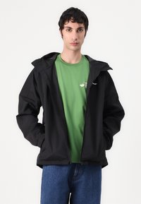 Black waterproof jacket with a hood, zip closure, and elastic cuffs, layered over a green t-shirt. Model stands against a plain background.