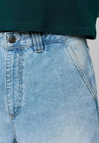 Shorts en denim bleu clair avec taille haute, dotés d'une fermeture à bouton à l'avant, de coutures visibles et d'une texture de tissu légèrement usé.