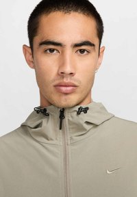 Beige jas met rits en een hoge kraag, verstelbare trekkoorden en een klein geborduurd logo op de borst. Gladde, lichte stof.