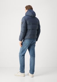 Temno modra puffer jakna s kapuco, prešita zasnova in logotip na rami; v kombinaciji s svetlo modrimi kavbojkami in belimi športnimi copati.