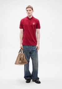 Κόκκινο πόλο μπλουζάκι με κεντημένο λογότυπο, συνδυασμένο με μπλε τζιν παντελόνι φαρδιάς γραμμής. Κρατώντας ένα καφέ δερμάτινο τσάντα, με δύο κοντά χερούλια.