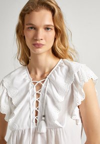 Blusa in cotone bianco con scollatura allacciata, maniche a volant in pizzo e un accent decorativo con frange. Tessuto liscio e vestibilità ampia.