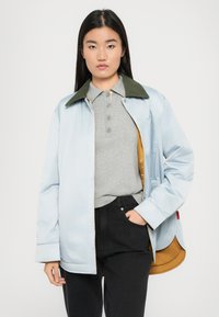 Lyseblå jakke med grønn krage, knappet front og gullfôr, båret over en grå polo-skjorte og svarte jeans.