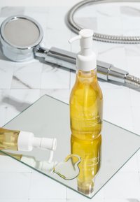Bottiglia trasparente di olio detergente giallo con tappo a pompa bianco, posizionata su una superficie specchiata accanto a un soffione cromato.