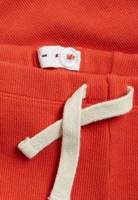 Oranssi puuvillakangas, jossa on valkoinen punottu naru. Vyötäröllä on valkoinen nappi, jossa on punainen ommel.