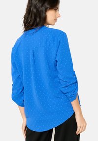 Blusa blu con una vestibilità rilassata, caratterizzata da un motivo a pois strutturato, colletto e maniche arrotolate. L'orlo è leggermente curvato sul retro.
