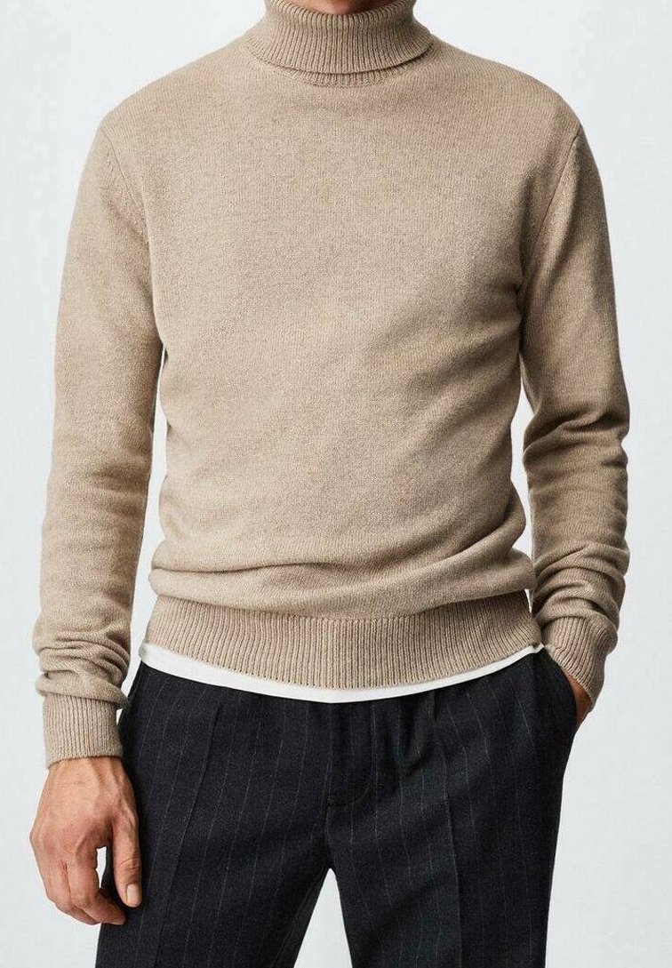 Beige Rollkragenpullover über einem weißen Hemd, kombiniert mit dunkel gestreiften Hosen, Hände entspannt an den Seiten.