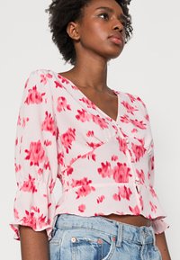 Blusa com estampado floral em rosa claro com detalhes em vermelho. Apresenta um decote em V, mangas bufantes com punhos franzidos e uma cintura ajustada.