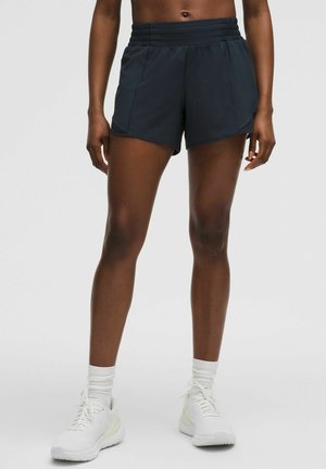 Vrouw draagt zwarte sportshorts, witte sokken en witte hardloopschoenen, staand tegen een effen lichte achtergrond.