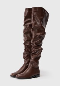 Botas marrones de cuero sobre la rodilla con un diseño holgado. Textura suave, punta redondeada y suela plana. Costuras decorativas a lo largo de los lados.
