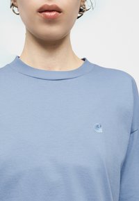 Άτομο που φοράει ένα φωτεινό γαλάζιο μπλουζάκι με στρογγυλή λαιμόκοψη και μικρό κεντητό λογότυπο κύματος στο αριστερό στήθος, με εμφανή τα χείλη και τον λαιμό.