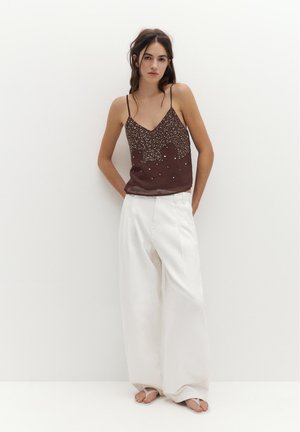 Młoda kobieta w brązowym topie bez rękawów z wzorem, szerokich białych spodniach i białych sandałach stoi oparta o białą ścianę z rękami za plecami.