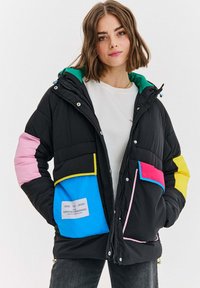 Žena v čiernej prešívanej bunde s farebnými nášivkami na vreckách a rukávoch, stojí s rukami v vreckách na bielom pozadí.