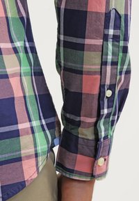Chemise à manches longues à carreaux verts, roses, bleu marine et beige, poignets boutonnés, portée avec un pantalon beige sur fond clair.