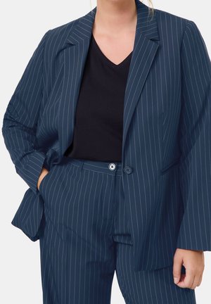Femme portant un costume à rayures bleu marine avec une chemise noire à col en V, mains dans les poches, debout devant un fond blanc uni.