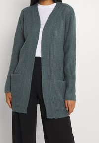 Šedý svetr s ribbed texturou, otevřený přední díl, dvě přední kapsy, dlouhé rukávy a volný střih. Nositelný přes bílou košili.