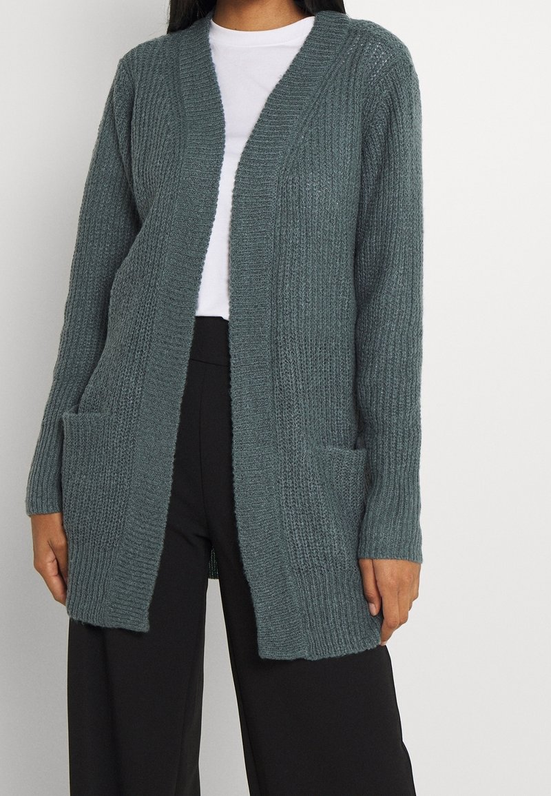 Šedý svetr s ribbed texturou, otevřený přední díl, dvě přední kapsy, dlouhé rukávy a volný střih. Nositelný přes bílou košili.