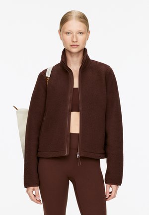 Brown fleece jacket with zip front, high collar, and relaxed fit. Paired with a brown crop top and leggings, featuring a beige bag.