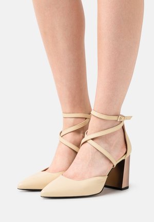 Pumps beige con cinturino alla caviglia, punta appuntita, cinghie incrociate e tacco a blocco affusolato con un accento metallico. Texture in pelle liscia.