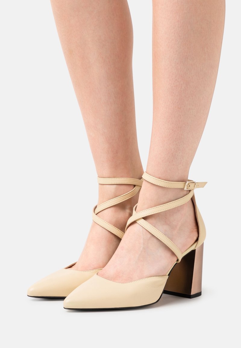 Béžové lodičky s pásem kolem kotníku, špičatou špičkou, křížovými popruhy a zúženým blokovým podpatkem s kovovým akcentem. Hladký kožený povrch.