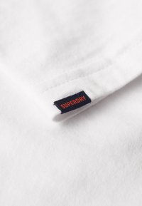 Weißes Baumwoll-T-Shirt mit einem schwarzen und orangen "SUPERDRY"-Label am Kragen, mit glatter Textur und klassischem Design.