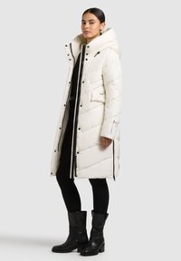Palton lung alb, matlasat, cu glugă mare, fermoar frontal și capse. Prezintă accente negre și detalii cu buzunare cu fermoar.