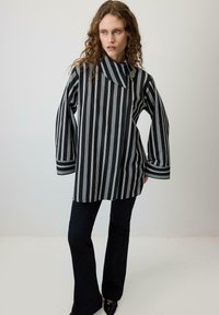 Ženska z dolgimi kodrastimi lasmi, oblečena v črno-bel suknjič z navpičnimi črtami in broško, črne široke hlače ter črne koničaste čevlje.