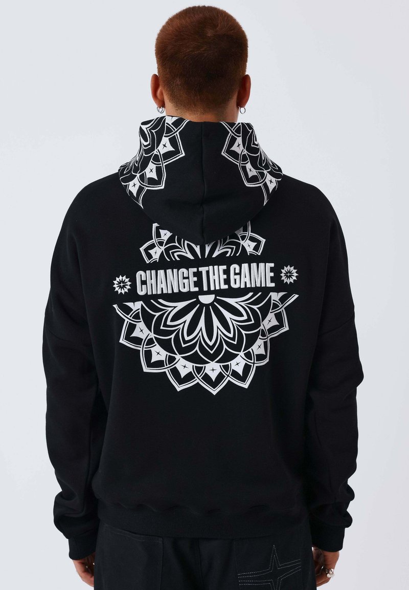 Personne aux cheveux courts portant un sweat à capuche noir orné d'un mandala floral blanc et du texte « CHANGE THE GAME » dans le dos, debout devant un fond uni.