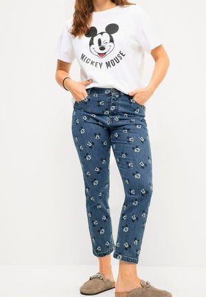 Femme portant un t-shirt blanc Mickey Mouse, un jean bleu à motifs de petits visages de Mickey Mouse et des mocassins beiges à enfiler.