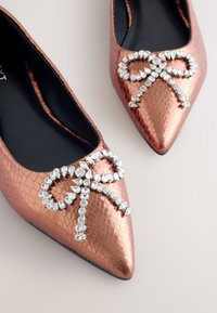 Ballerines couleur cuivre à bout pointu avec une finition vernie texturée. Ornées d'un nœud en strass sur le dessus. Semelle intérieure noire.
