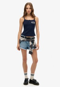 Navy tanktop met kantafwerking en print "BOSTON SUPERDRY", gecombineerd met denim shorts, een grijs sweatshirt om de taille geknoopt, en zwarte schoenen.