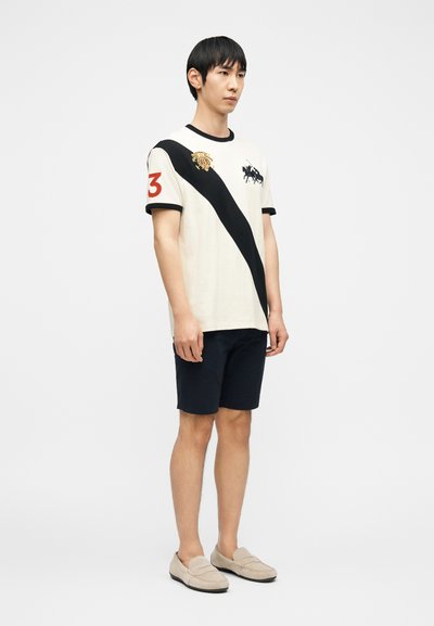 Νεαρός άνδρας που στέκεται, φορώντας λευκό μπλουζάκι με μαύρη διαγώνια ρίγα, κόκκινο αριθμό 3 στο μανίκι, μαύρο σορτς και μπεζ μοκασίνια.