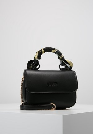 Schwarze Liu Jo Handtasche mit einem obersten Griff, der mit Blumenstoff umwickelt ist, und einem goldenen Kettengurt, die auf einer weißen Oberfläche liegt.