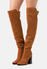 Stivali over-the-knee in suede marrone con borchie argentate lungo i lati, tacco a blocco e zip sul lato interno. Texture liscia.