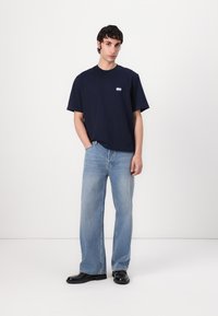 Navy blue cotton t-shirt with short sleeves, featuring a small blue and white logo. Paired with light blue wide-leg jeans and black shoes.