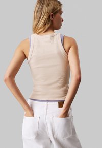 Top in cotone beige a coste sovrapposto a una canotta lilla chiara, abbinato a pantaloni in denim bianchi con tasche posteriori e un'etichetta in pelle.