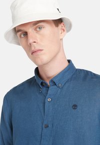 Camisa azul de botones hecha de lino con un acabado texturizado y botones oscuros; presenta un pequeño logo en el lado izquierdo del pecho. Sombrero tipo bucket blanco.