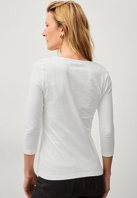 Bílé dlouhé tričko s dlouhým rukávem a kulatým výstřihem, přiléhavého střihu a hladké, měkké textury. Má klasický design bez ozdob.